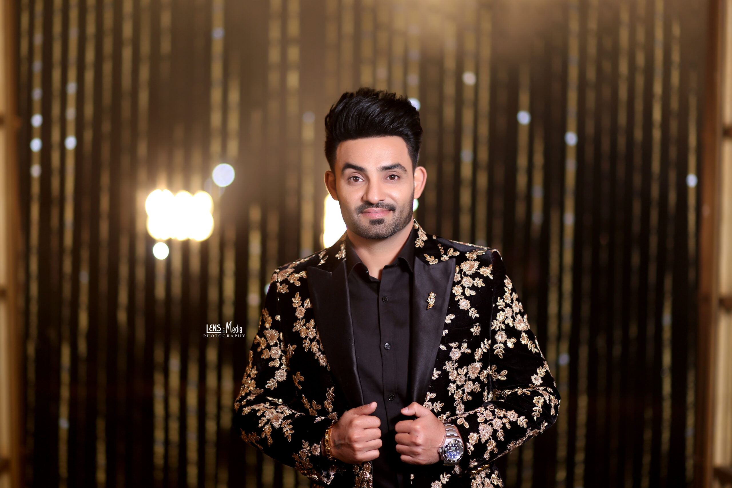 Resham singh Anmol wearing a black embroidered blazer posing against a golden light background